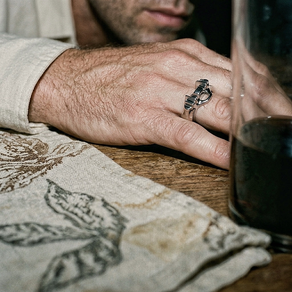 Sculptural sterling silver ring on male hand resting beside wine glass and linen napkin, editorial warm light