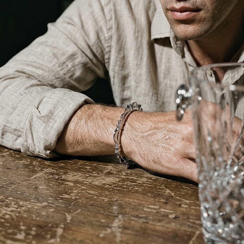Sterling silver bangle on male forearm in warm editorial light, with linen shirt cuff and crystal glass on dark wooden bar